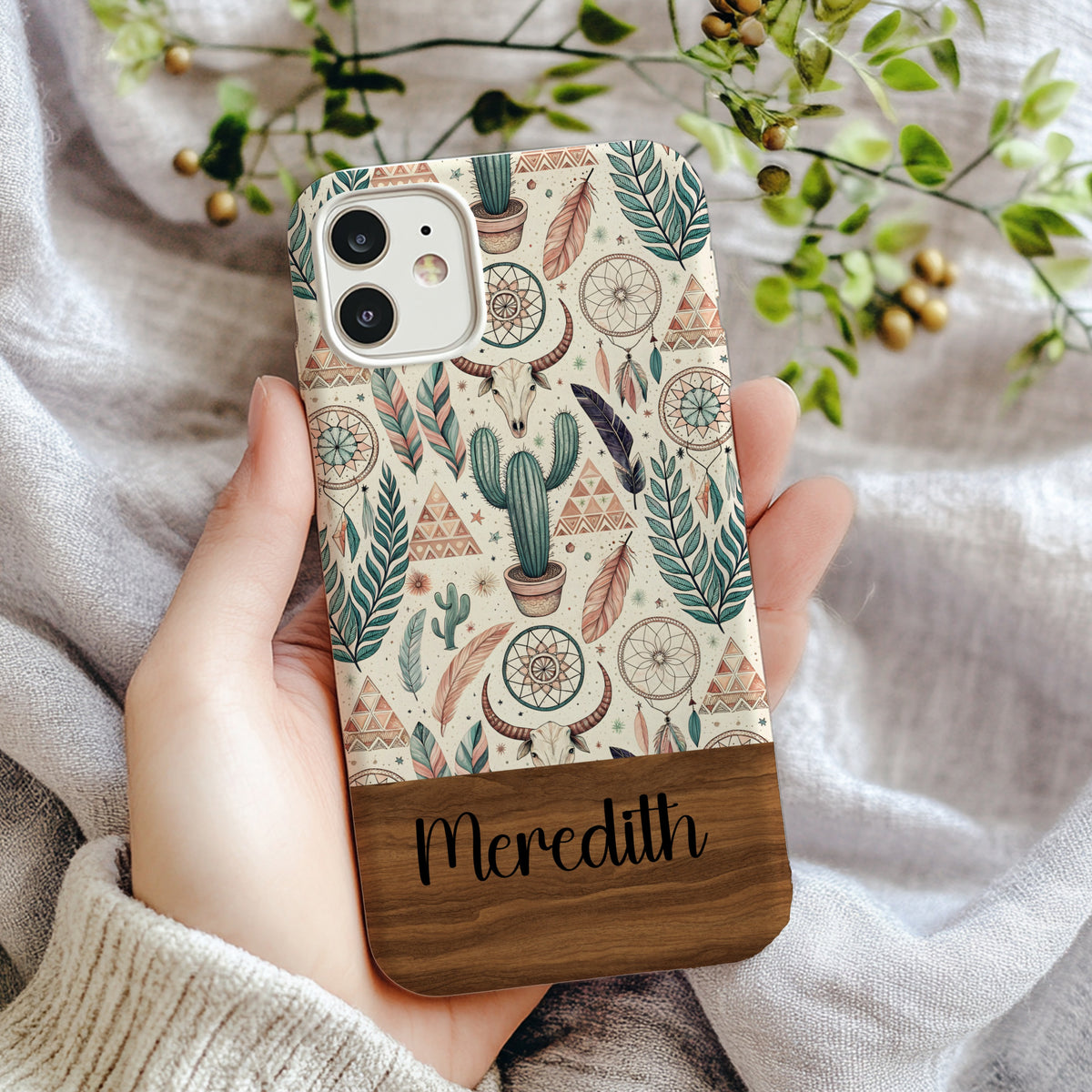 Boho-style iPhone case held in hand featuring a repeating pattern of cacti, dreamcatchers, feathers, and a bull skull in teal and brown on a cream background, with a wood-grain nameplate reading 'Meredith'
