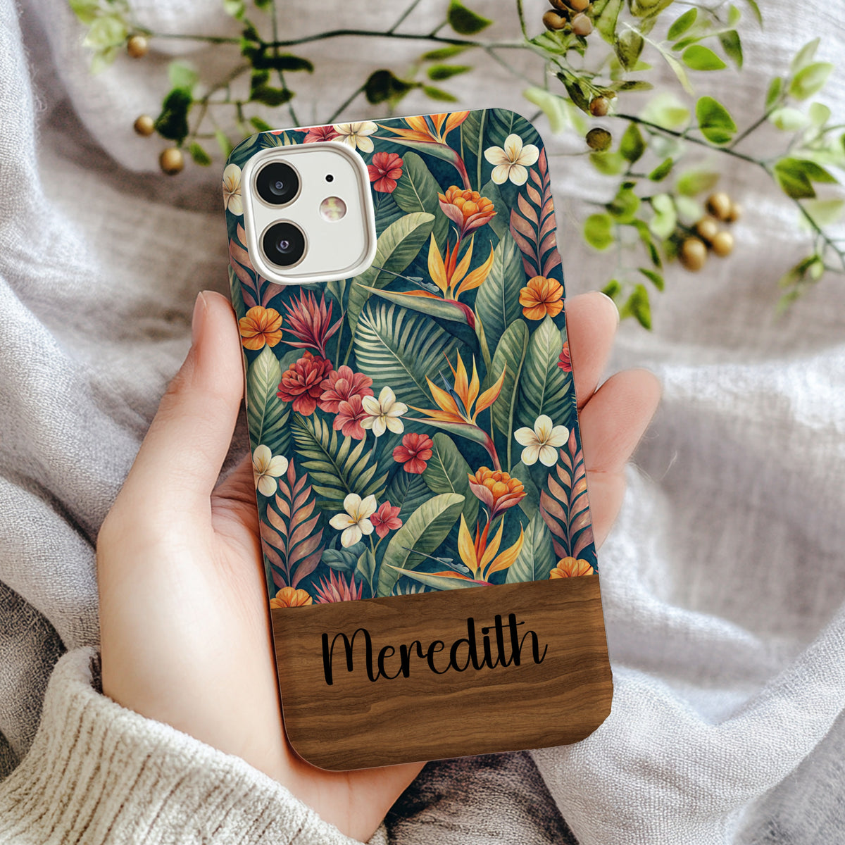 Dark teal phone case with watercolor bird of paradise, red hibiscus, and white plumeria flowers among green palm leaves, personalized with a custom name in black cursive script on a dark wood bottom band.