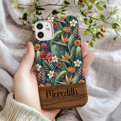 Dark teal phone case with watercolor bird of paradise, red hibiscus, and white plumeria flowers among green palm leaves, personalized with a custom name in black cursive script on a dark wood bottom band.
