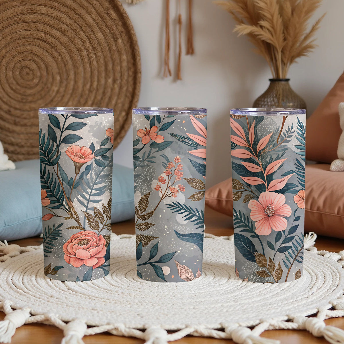 Three 20oz stainless steel skinny tumblers with clear lids printed with a grey-toned floral pattern featuring pink and peach flowers, gold leaves, and teal botanical foliage, displayed on a woven cream rug in a boho-style living room.