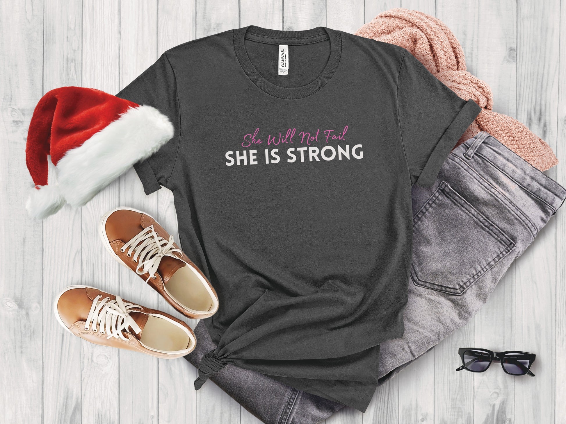 Dark grey t-shirt with "She Will Not Fail SHE IS STRONG" text, flat lay with Santa hat, jeans, sweater, and shoes.