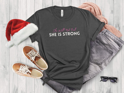 Dark grey t-shirt with "She Will Not Fail SHE IS STRONG" text, flat lay with Santa hat, jeans, sweater, and shoes.