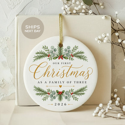 Round ceramic ornament reads "Our First Christmas As A Family of Three" and "2026".